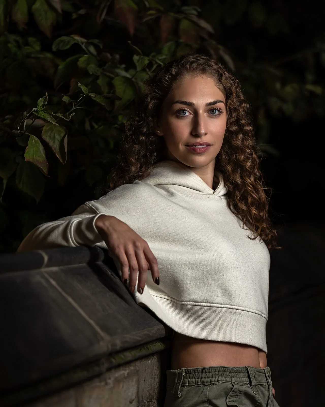 Night portrait of a curly-haired woman in a cream hoodie against dark garden greenery, New Haven area